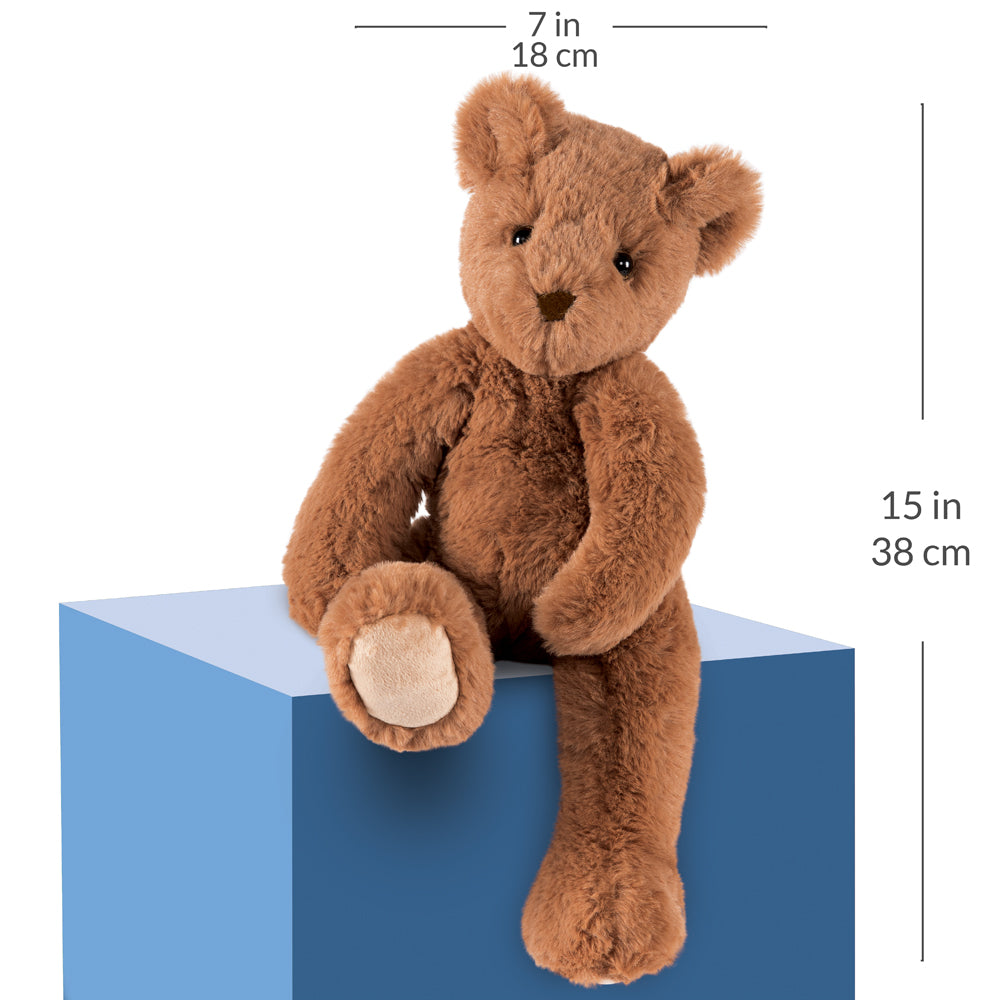 Brown plush teddy bear sitting on a blue display block with dimension lines and text showing 7 in / 18 cm width and 15 in / 38 cm height on a white background.