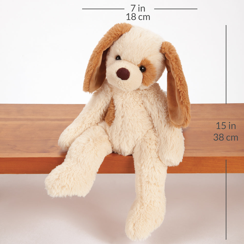 A plush dog toy with cream-colored fur and brown ears, sitting on a wooden shelf. Measurement lines and text indicate dimensions of 7 in / 18 cm width, 15 in / 38 cm height, and the overall height of 18 in / 46 cm.