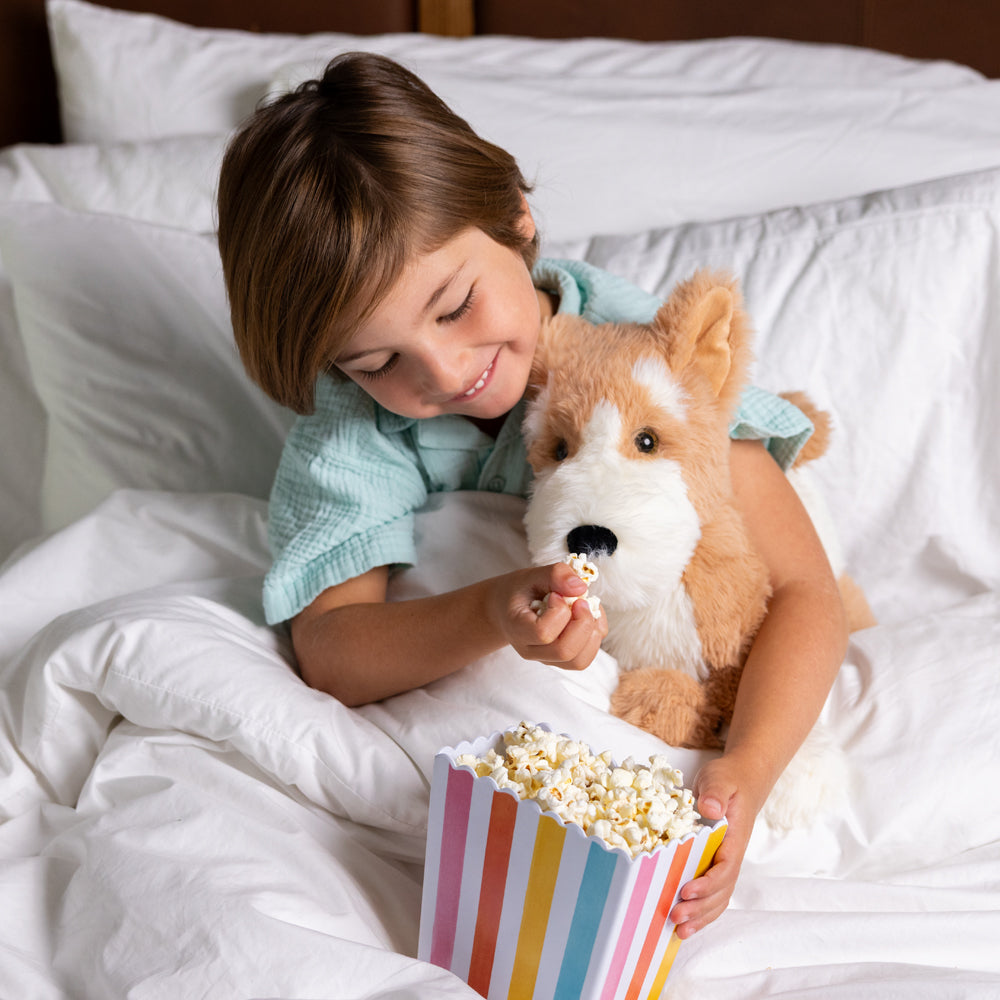 A young person in bed, smiling as they feed popcorn to a plush corgi dog stuffed animal. 