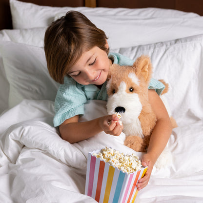 A young person in bed, smiling as they feed popcorn to a plush corgi dog stuffed animal. 