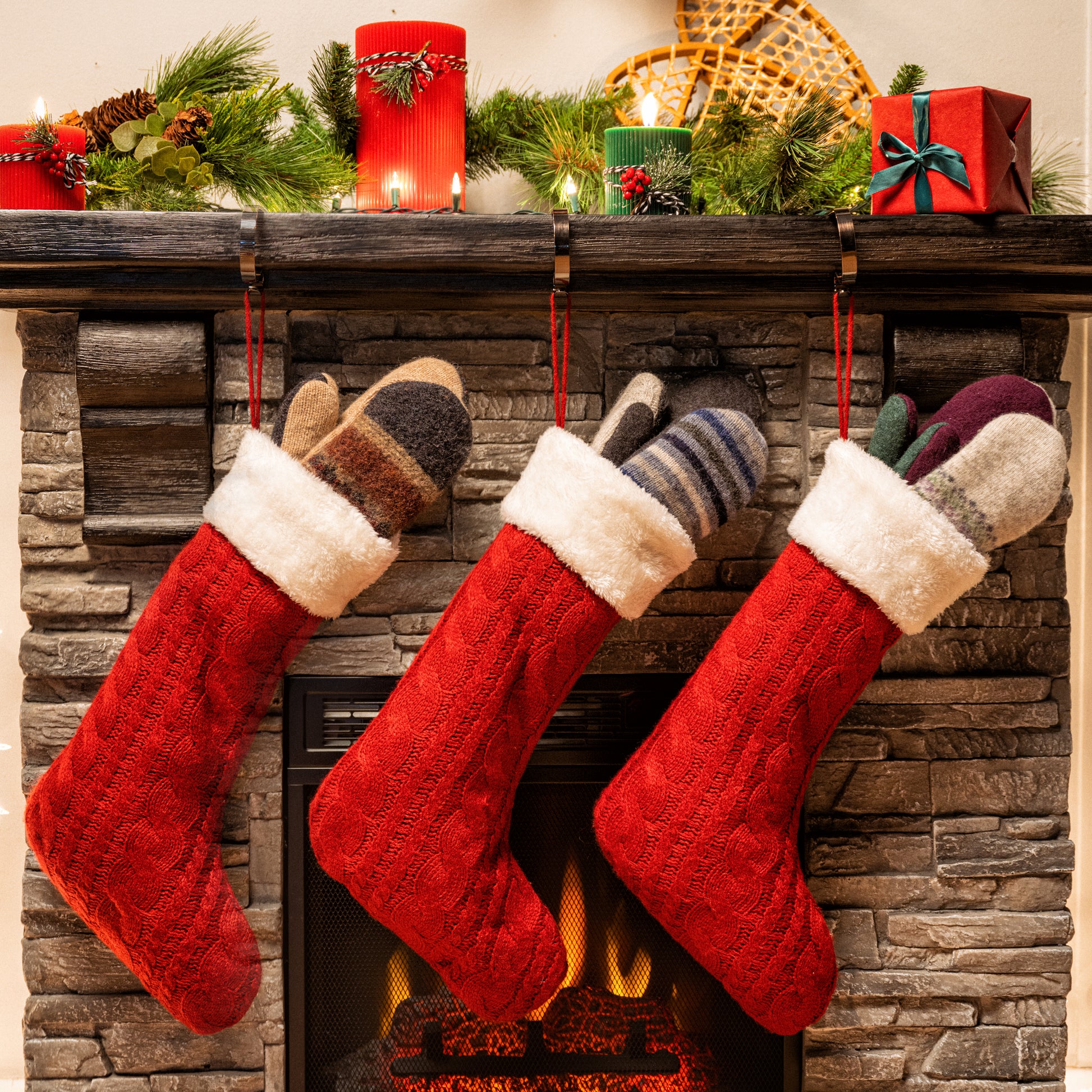 Red Christmas stockings hanging on a fireplace mantle with candles and decorations.