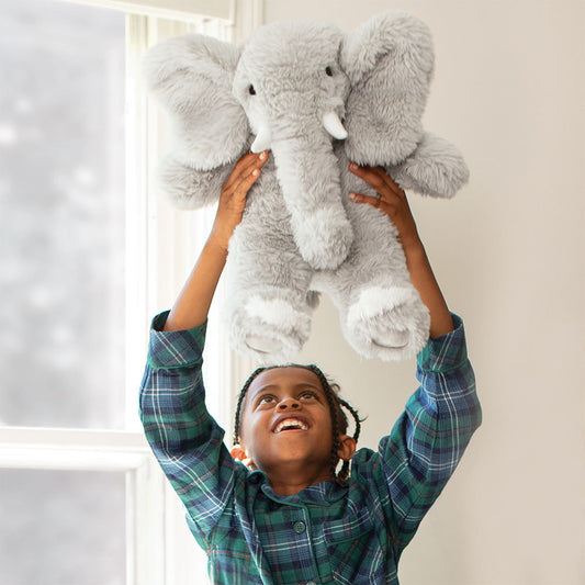 Child holding a large soft gray elephant plush toy with white tusks and feet in bright room.