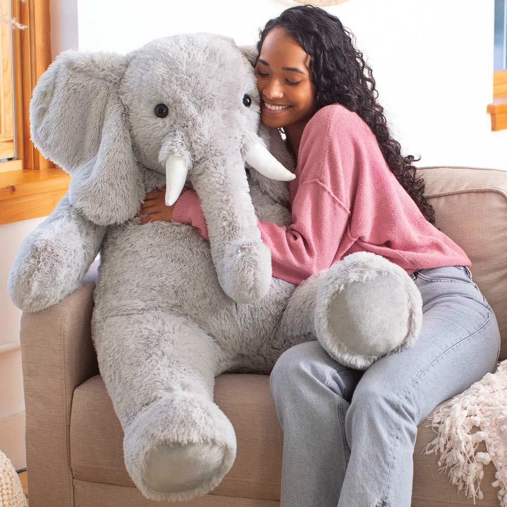 Large gray plush elephant with big ears and white tusks sitting on beige couch with woman in pink sweatshirt and jeans.