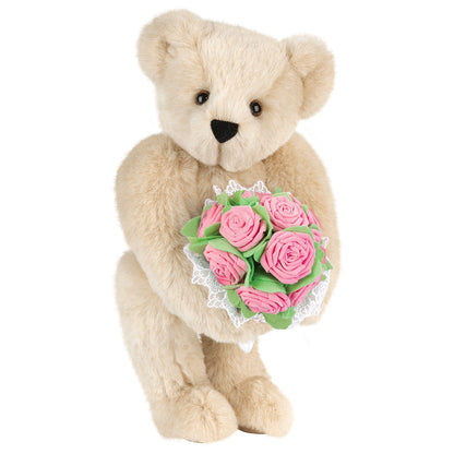 Cream-colored teddy bear holding a bouquet of pink rose flowers with green leaves and white lace trim.