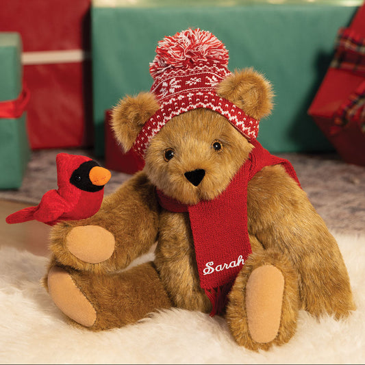 Honey-colored plush bear wearing a red knit hat with white patterns and a red scarf, holding a red cardinal bird plush.